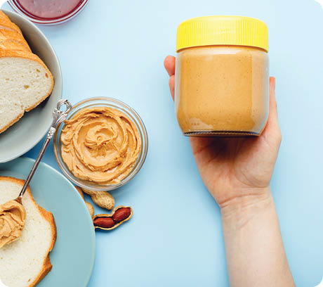 Creamy peanut paste in glass jar with yellow cap in female hand and peanuts in the peel scattered on blue colored background  Cooking breakfast with peanut butter  Vegan food concept 